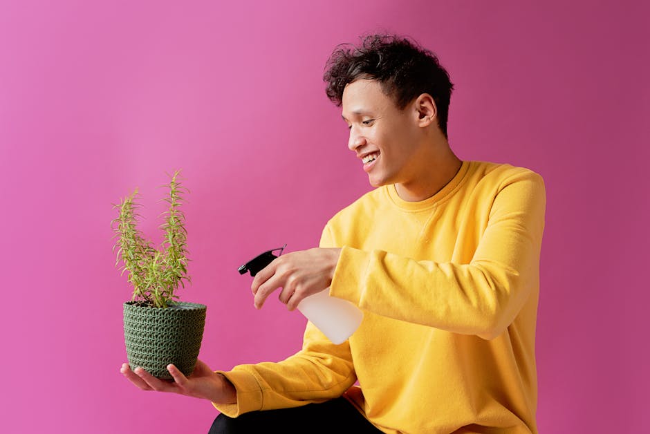 Young man smiling while watering a potted plant with a spray bottle against vibrant pink background.