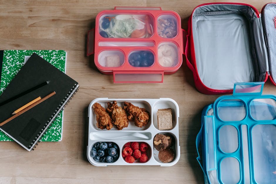Overhead view of healthy lunchboxes with fruits, chicken, and notebooks on a wooden desk.