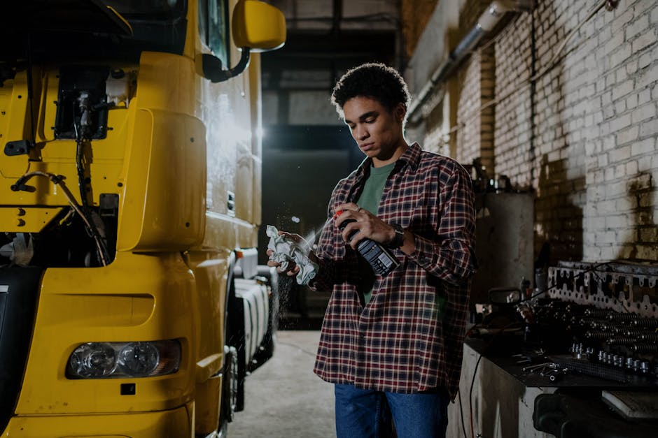 A mechanic in a plaid shirt sprays lubricant while repairing a truck indoors.