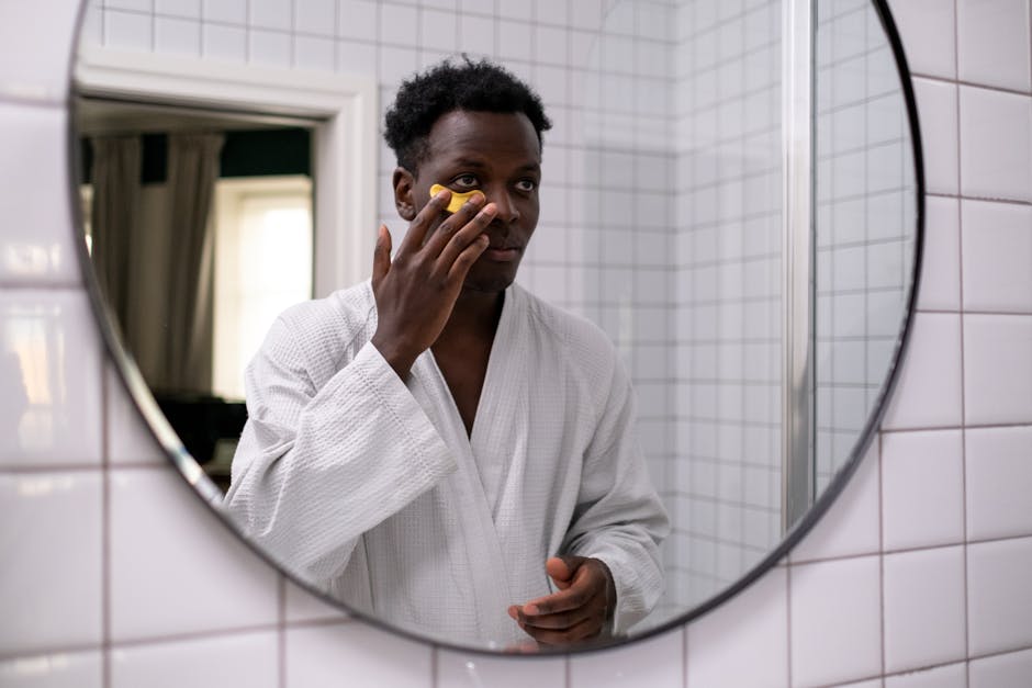 A man wearing a bathrobe applies an under-eye patch in front of a round mirror in a tiled bathroom.