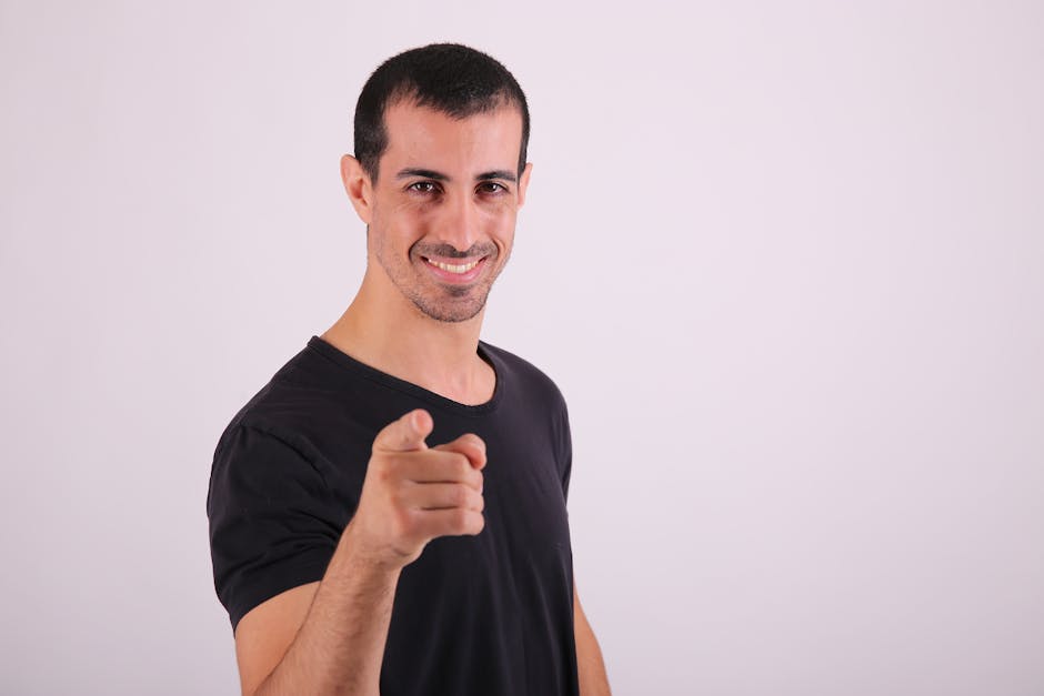 A confident young man in a black shirt points playfully at the camera in a studio setting.