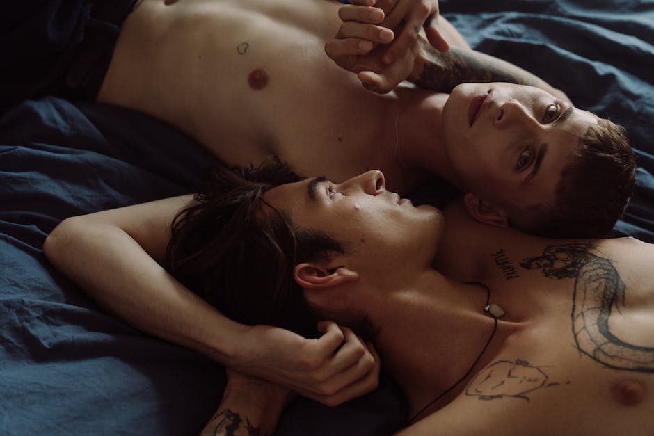 Two tattooed men embracing intimately in a bedroom setting.