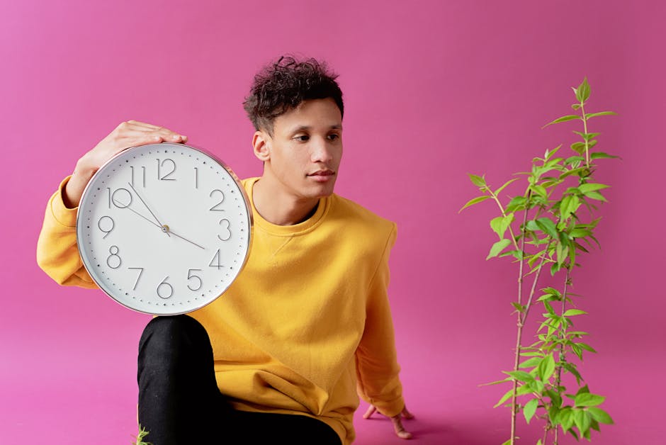 A young man sitting with a clock and green plant against a pink studio backdrop.