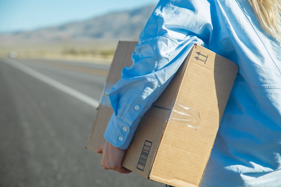 A woman holds a cardboard box while walking on a sunny road, wearing a blue shirt.
