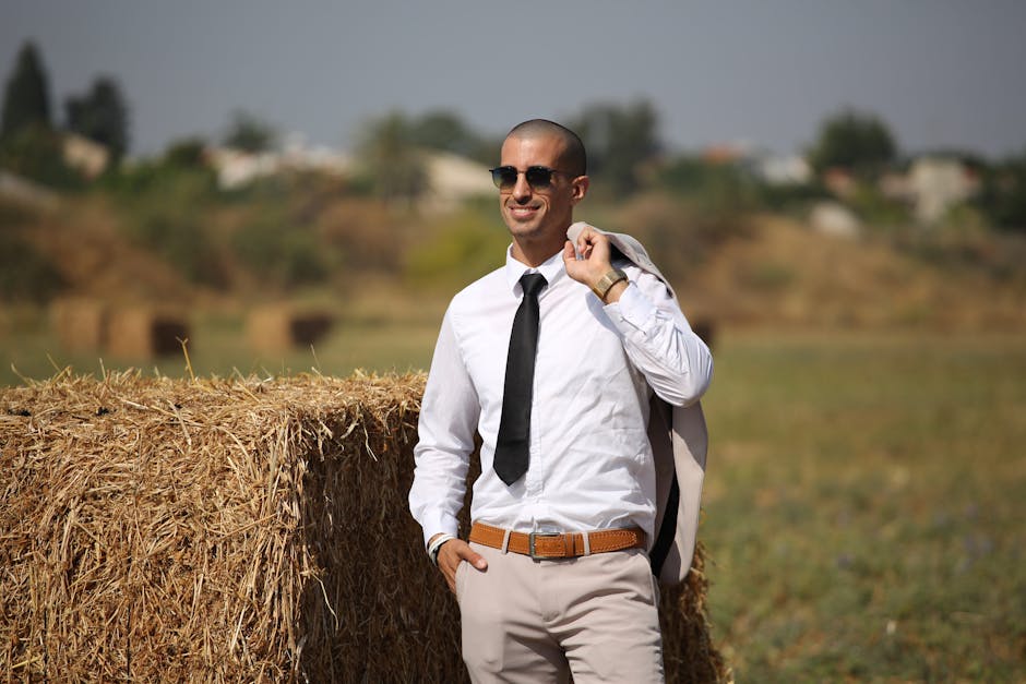 Confident man in sunglasses with suit jacket, smiling in a hay field.