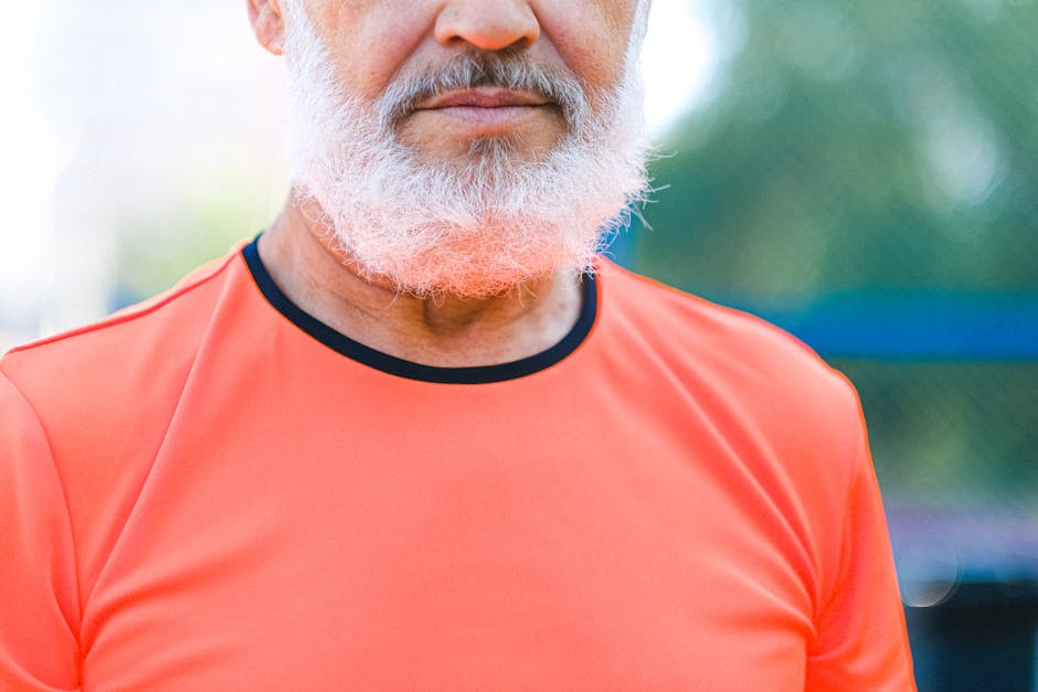 Close-up of a mature man with a white beard wearing a sporty orange shirt outdoors.