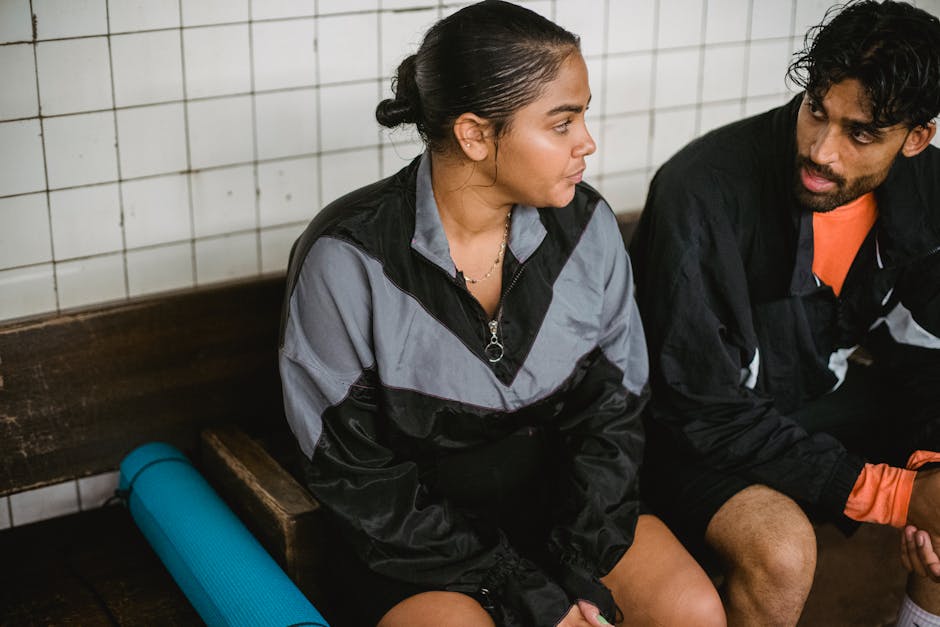 A man and woman in sportswear relax on a bench indoors post-workout, enjoying some downtime.