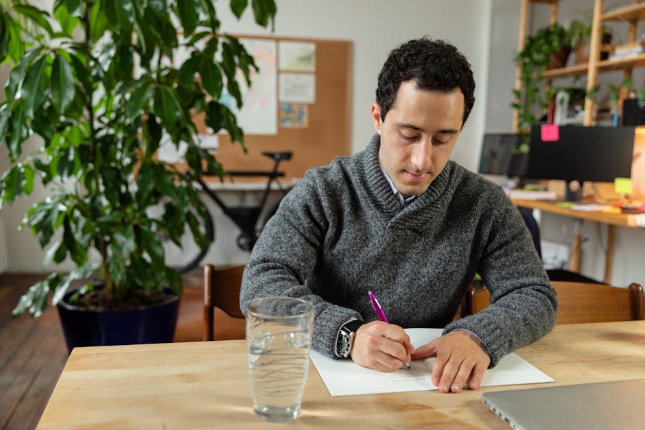 Man in a gray sweater writing at a desk in a cozy office with plants.